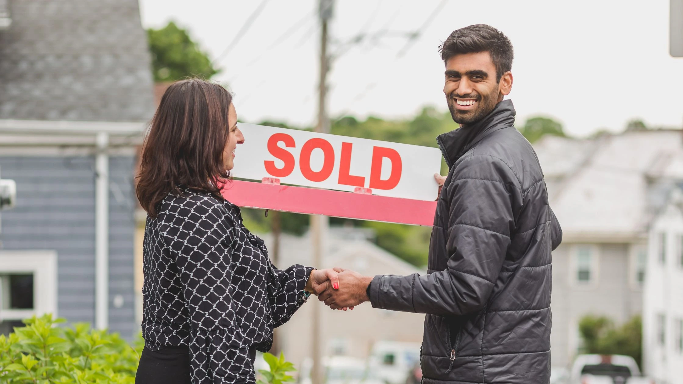 Two people shaking hands in front of a home while holding a “SOLD” sign, illustrating a successful sale for a Real Estate Selling Guide.