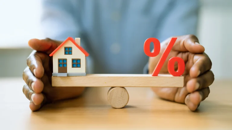 A person holds a wooden balance board with a small model house on one side and a red percentage symbol on the other, symbolizing how interest rate cuts impact home affordability in Canada’s 2025 housing market.